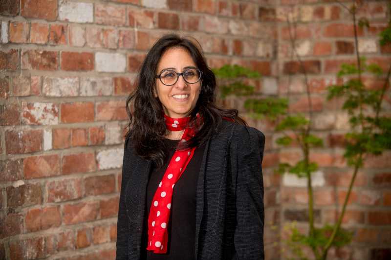 woman with long dark hair and dark-rimmed glasses standing against a red brick wall